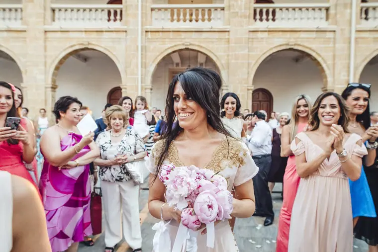 Laughing bride holding pink peonies among clapping female guests in the courtyard