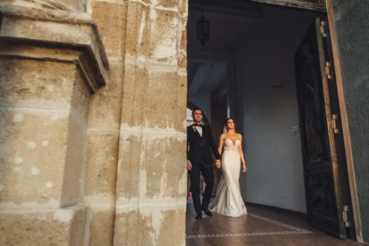 Bride and groom stepping out of a tall wooden church doorway at dusk