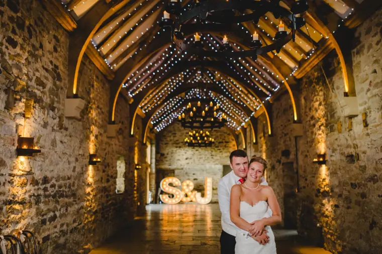 Couple standing close in the fairy-lit stone hall with S&J letters glowing behind