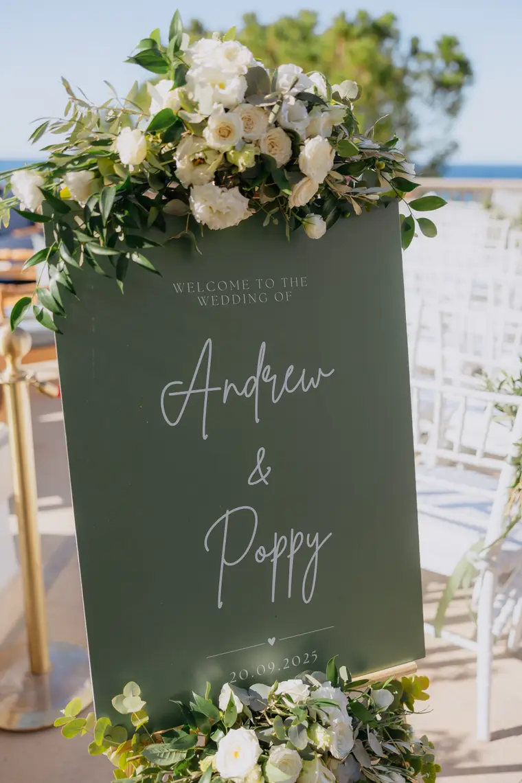 Green welcome sign reading Andrew & Poppy, trimmed with white flowers