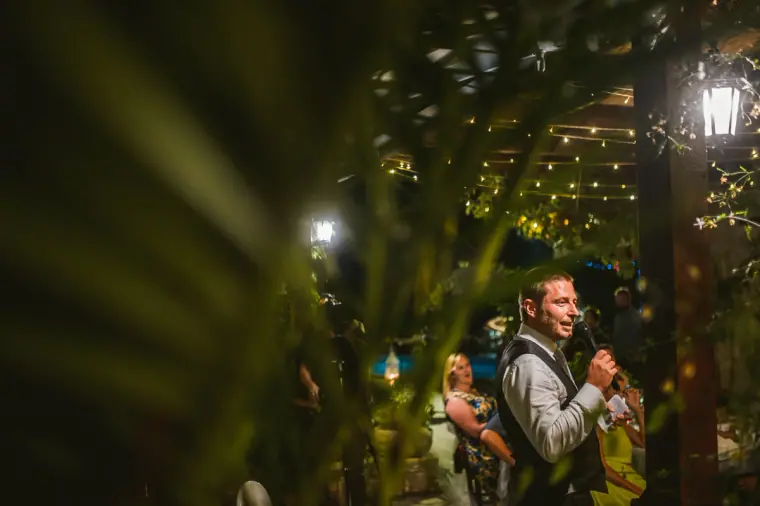 Groom giving his speech into a microphone, framed by leaves and lanterns
