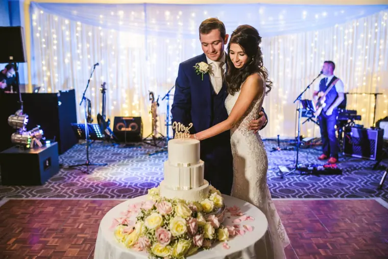 Elina and Iain cutting their tiered wedding cake as the band plays behind them