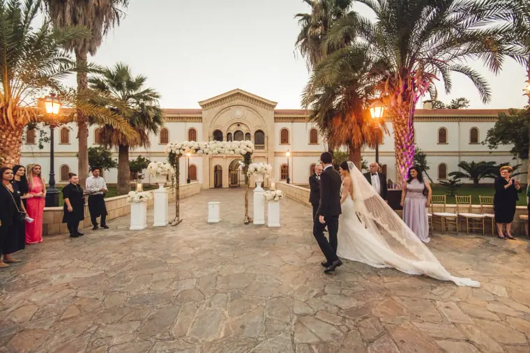 Bride and groom crossing a palm-lined courtyard towards a grand building at sunset