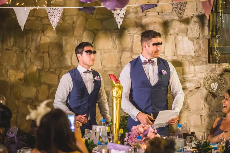 Two groomsmen in waistcoats giving a speech, one in sunglasses, beneath bunting