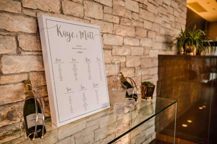 Kaye and Matt's table plan on a glass shelf against a stone wall, with Champagne