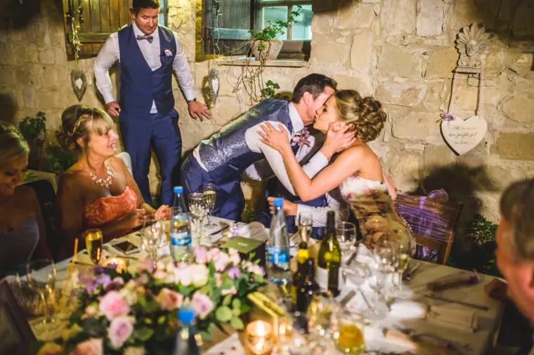 Groom leaning in to kiss the bride at the top table as the best man stands by