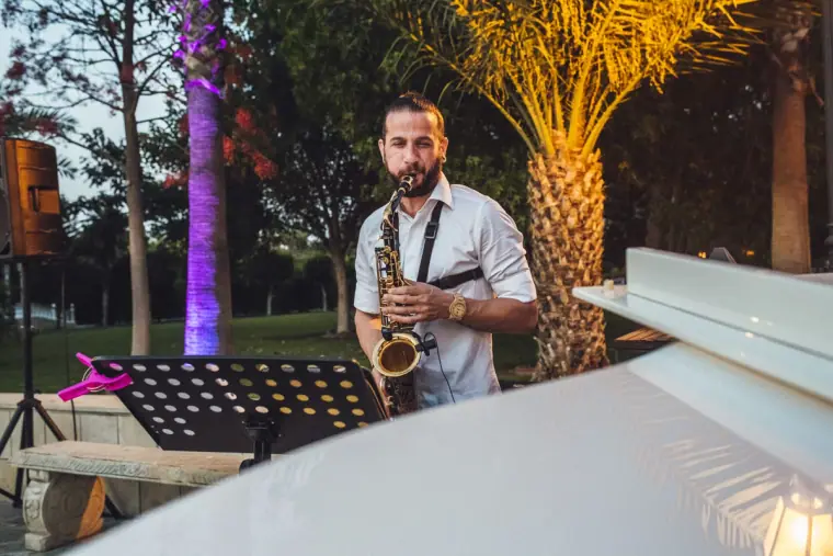 Saxophonist performing beside a white grand piano under lit palm trees