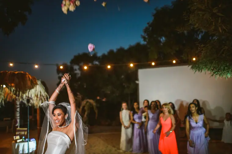 Bride raising her arms in celebration as bridesmaids in lilac look on at dusk