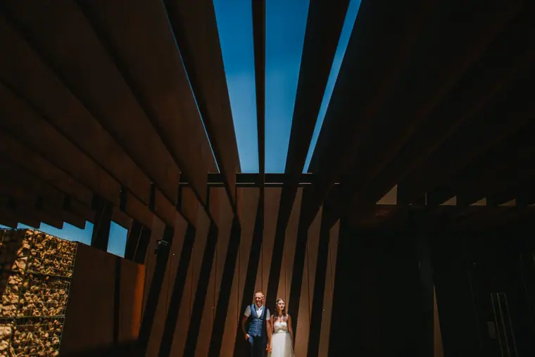 Sarah and Martin standing small beneath dramatic slatted roof shadows at Nissi Beach, Ayia Napa