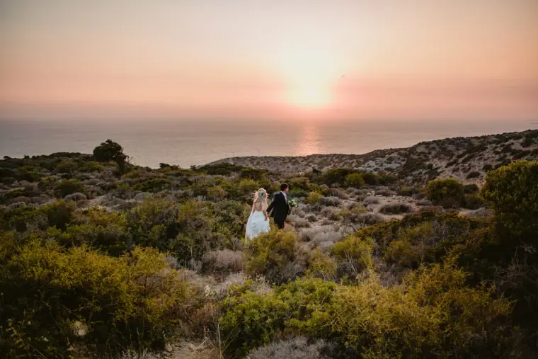 Couple standing on a scrubby headland as the sun sets over the sea