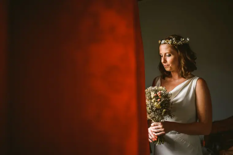 Fleur in a satin gown and flower crown holding her bouquet beside a red curtain, Vasilias Nikoklis Inn