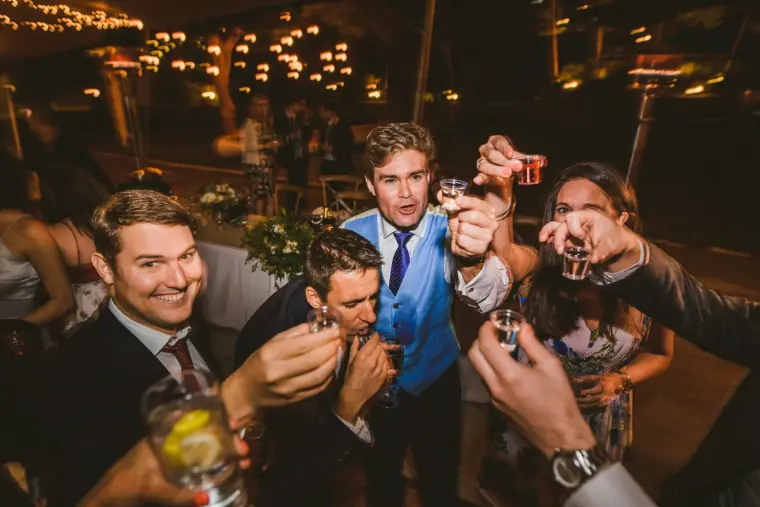 Groom and guests raising shot glasses together under festoon lights