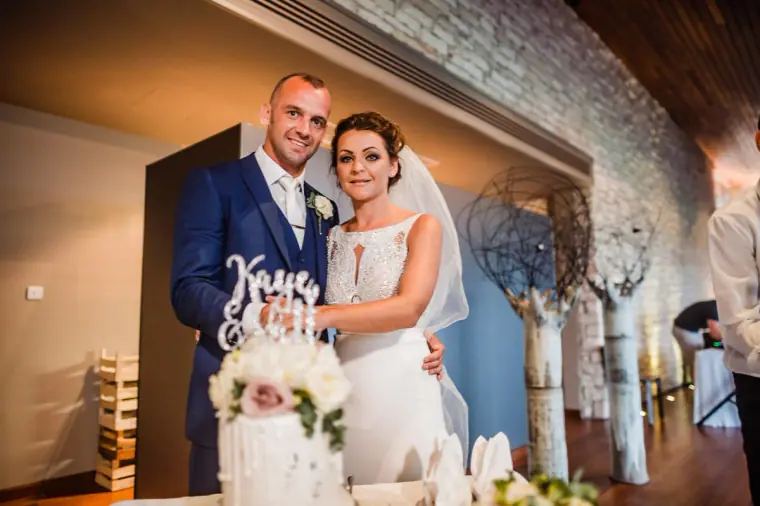 Bride and groom posing behind their white cake with a "Kaye & Matt" topper