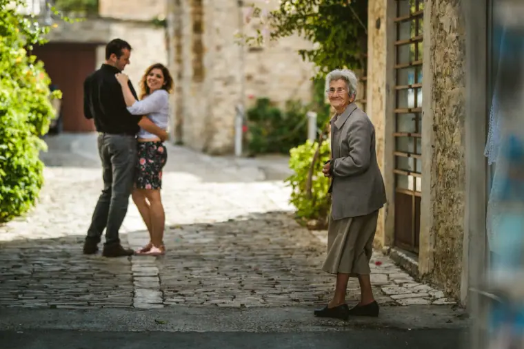 Nermine and Chris embracing on a cobbled lane as an elderly local looks on, Lefkara