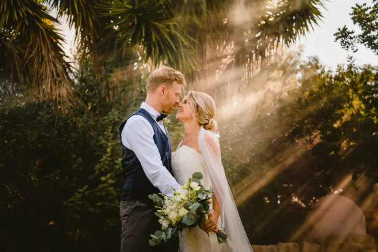 Groom kissing the bride's forehead as sunbeams filter through palms