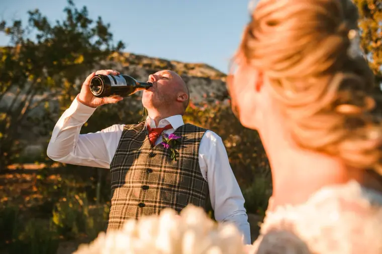 Groom in a tweed waistcoat swigging from a bottle as his bride looks on, Secret Valley, Paphos