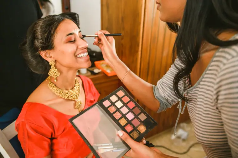 Bride in a red sari and gold jewellery laughing as her make-up is applied