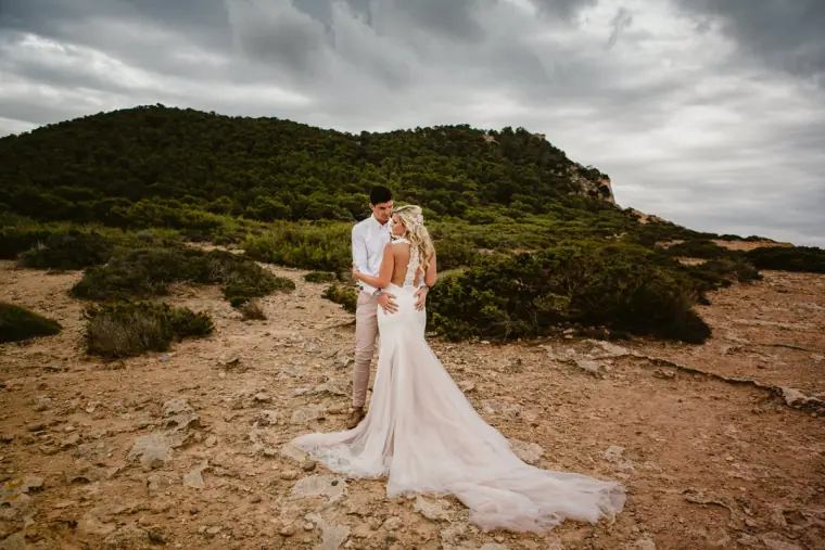 Bride and groom embracing on a rugged hillside under a cloudy sky, her train spread on the ground