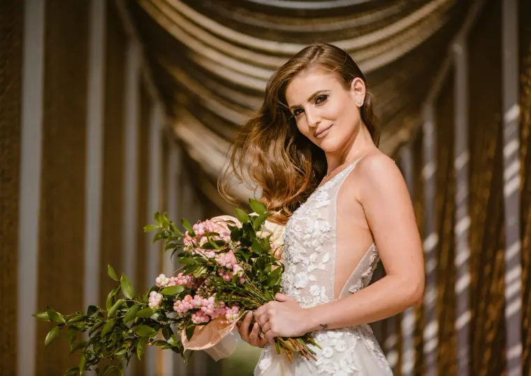 Bride in a plunging lace gown holding a pink bouquet beneath draped fabric