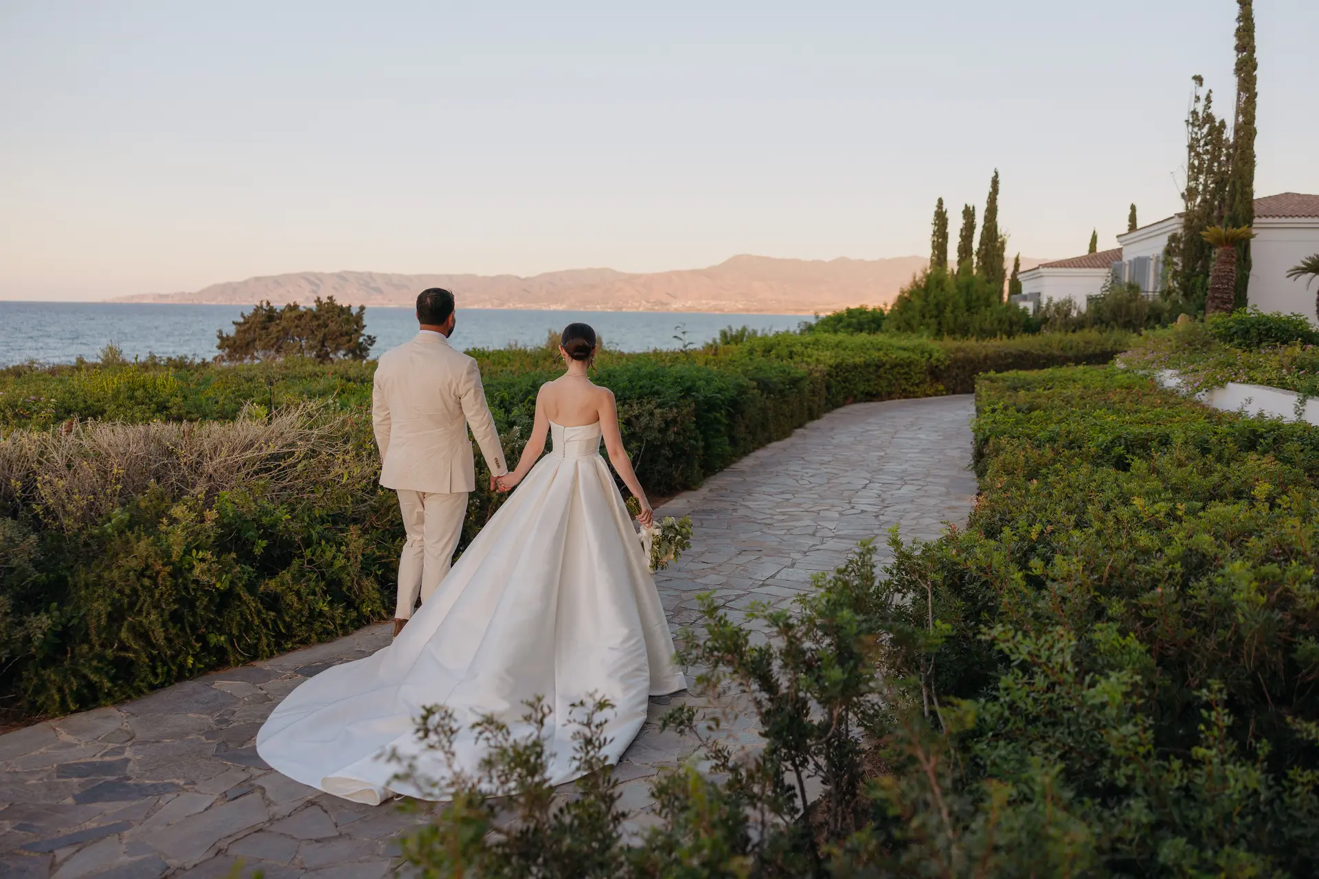 Bride and groom walking hand in hand along a garden path towards the sea