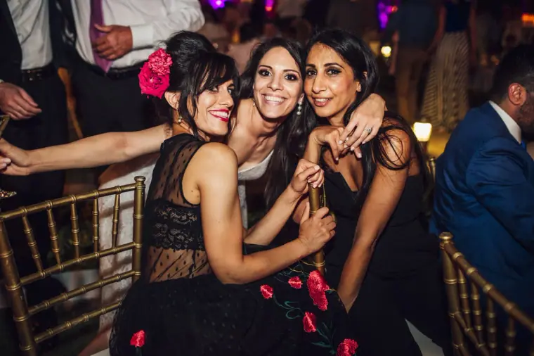 Bride hugging two friends on the dance floor, one wearing a red rose in her hair