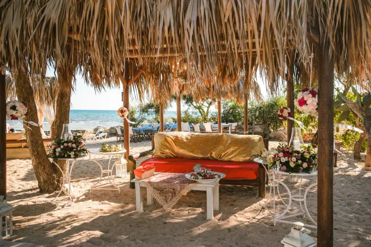Low red-cushioned seating under a thatched palm canopy on the beach