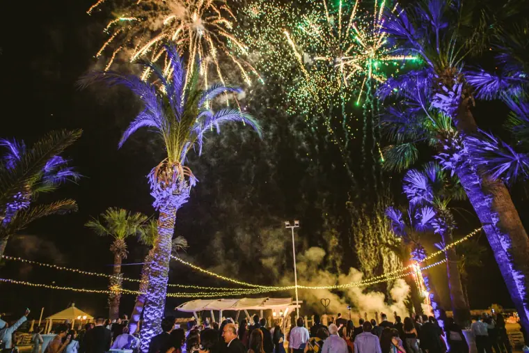 Fireworks bursting above palm trees lit in purple at an outdoor wedding reception