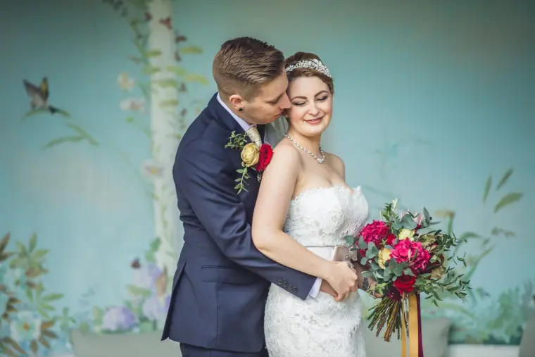 Charlie embracing Sarah from behind as she holds her red bouquet