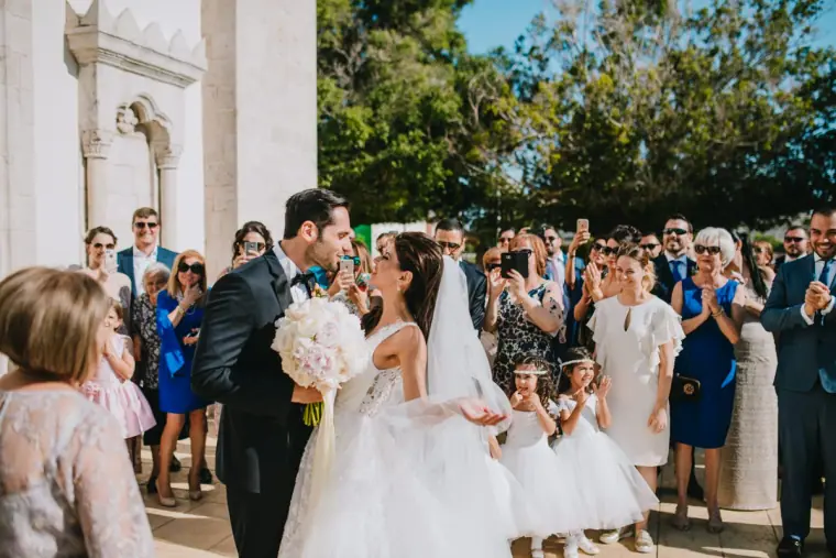 Couple sharing a kiss as guests and flower girls look on outside a white building, Limassol