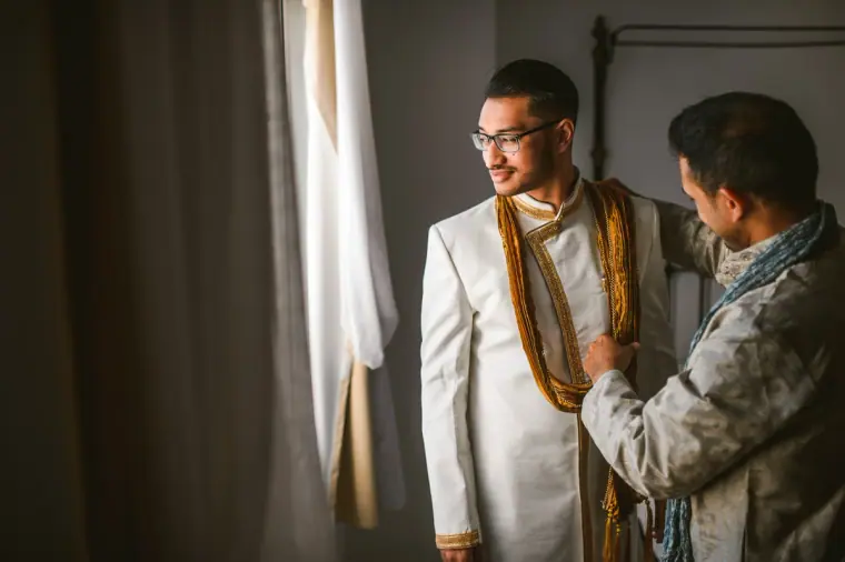 Older relative adjusting the groom's gold scarf by the bedroom window