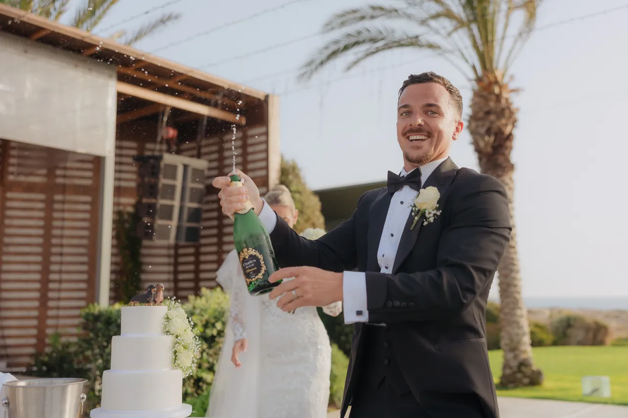 Groom grinning as he pops open a bottle of champagne