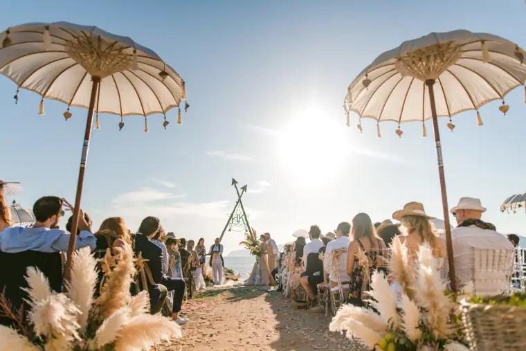 Beach ceremony with guests under fringed parasols and pampas grass lining the aisle to a wooden arch