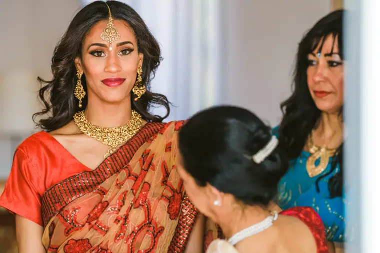 Raya in a red sari and gold maang tikka as relatives help fasten her outfit