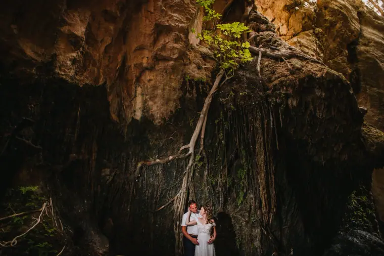 Kimberley and Craig dwarfed by a huge rock overhang draped in roots, Akamas