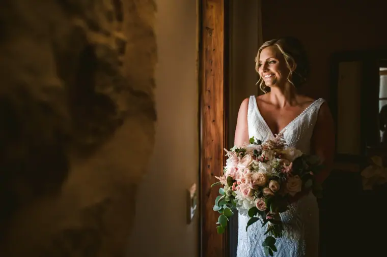 Bride in a V-neck gown holding a blush bouquet by a doorway in soft window light