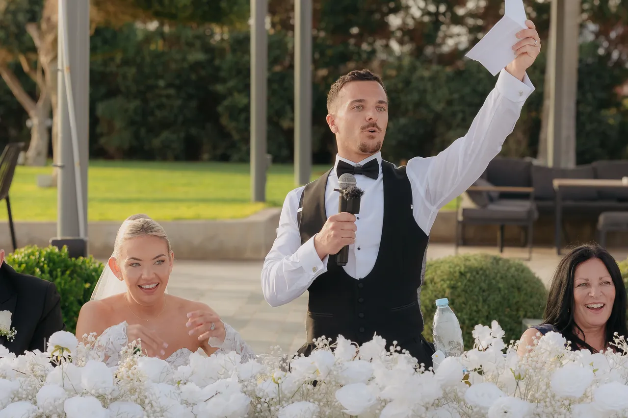 Groom waving a napkin mid-speech as his bride laughs beside him