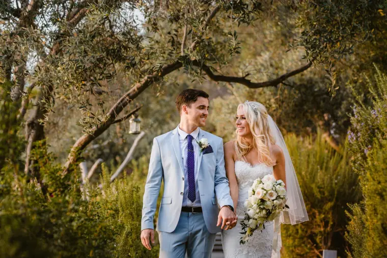 Groom in a pale blue suit and bride with her bouquet walking hand in hand under olive trees