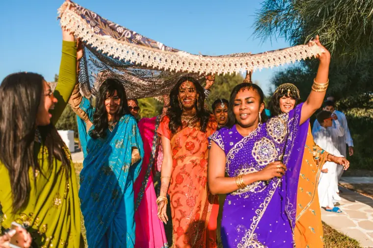 Bride walking beneath a canopy held aloft by women in jewel-coloured saris
