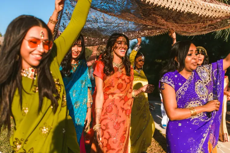 Smiling women in green, purple and orange saris raising the canopy over the bride
