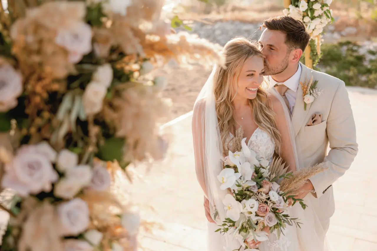 Dan kissing Becky's cheek beside a blush floral arrangement