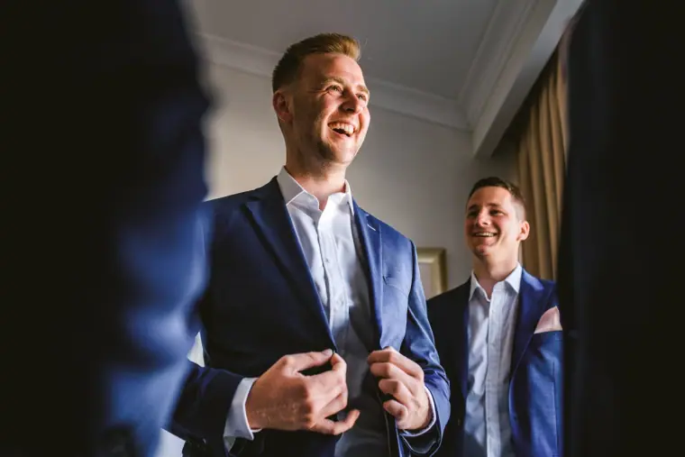Laughing groom buttoning his navy jacket as a groomsman watches