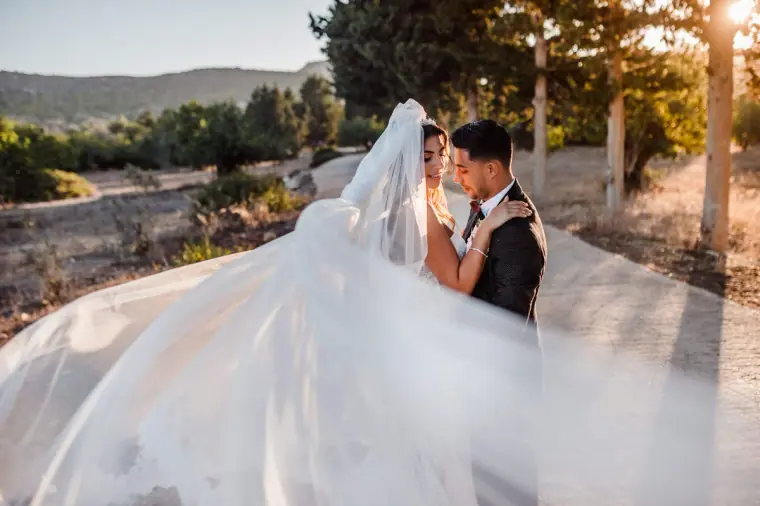 Bride's long veil sweeping across the frame as the couple kiss on a path, Anassa