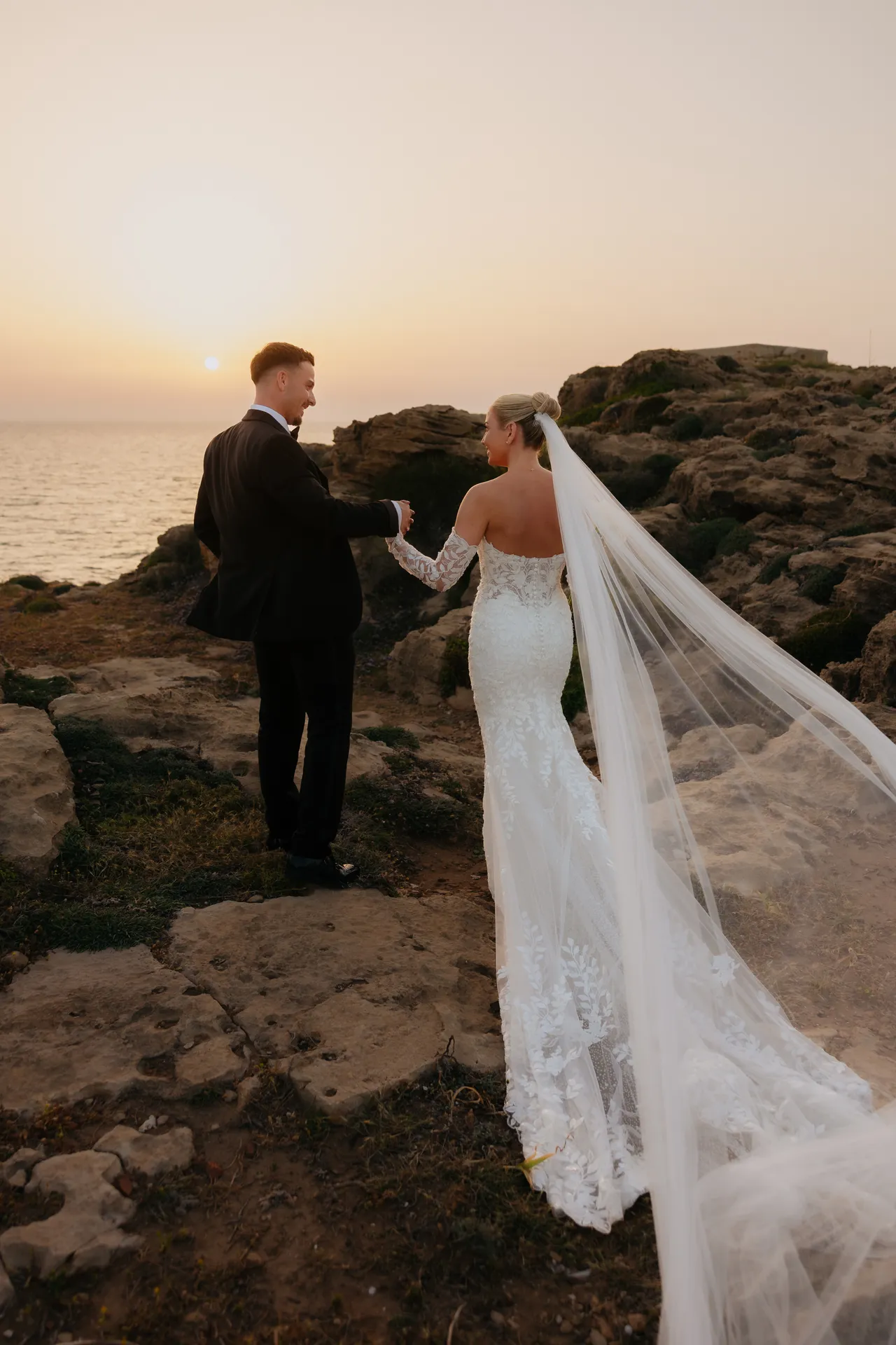 Groom leading his bride over the rocks at sunset, her veil blowing