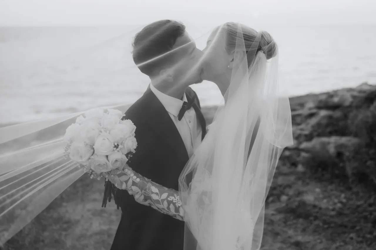 Black-and-white photo of the couple kissing as her veil blows in the wind