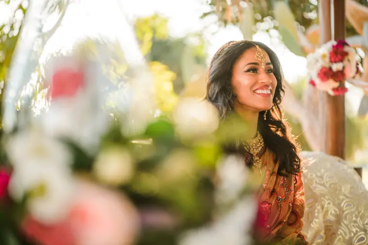 Bride smiling beneath the thatched canopy, framed by blurred flowers