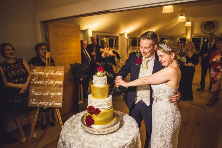 Sarah and Charlie cutting their gold and white wedding cake as guests watch