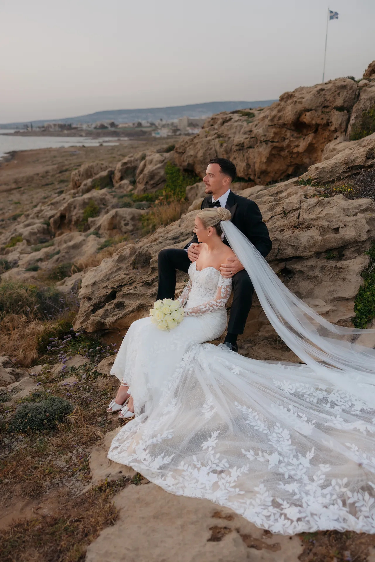 Bride and groom seated on the rocks above the sea at sunset