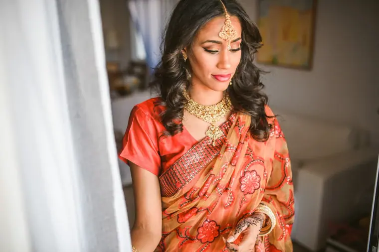 Raya in a coral sari, gold necklace and maang tikka by a sunlit window, Ayia Napa
