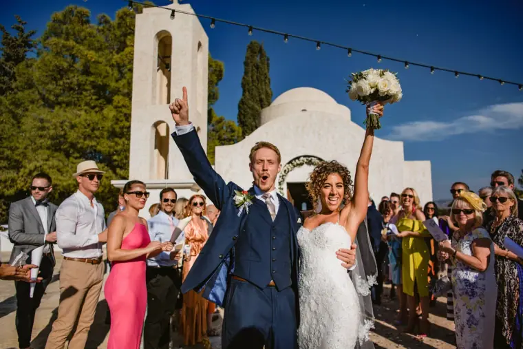 Groom punching the air and bride raising her bouquet outside a white chapel as guests cheer