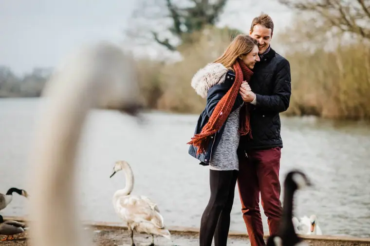 Couple cuddling in winter coats by a lake with swans during an engagement shoot